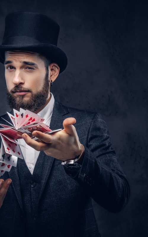 Portrait of a handsome magician in a black suit,  showing trick with playing cards on a dark background.